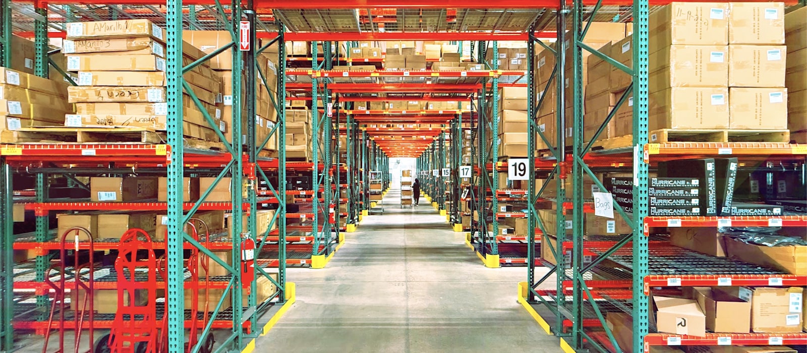 Numbered aisles of pallet racking in a wholesale distribution warehouse, hand trucks parked at the end of a row